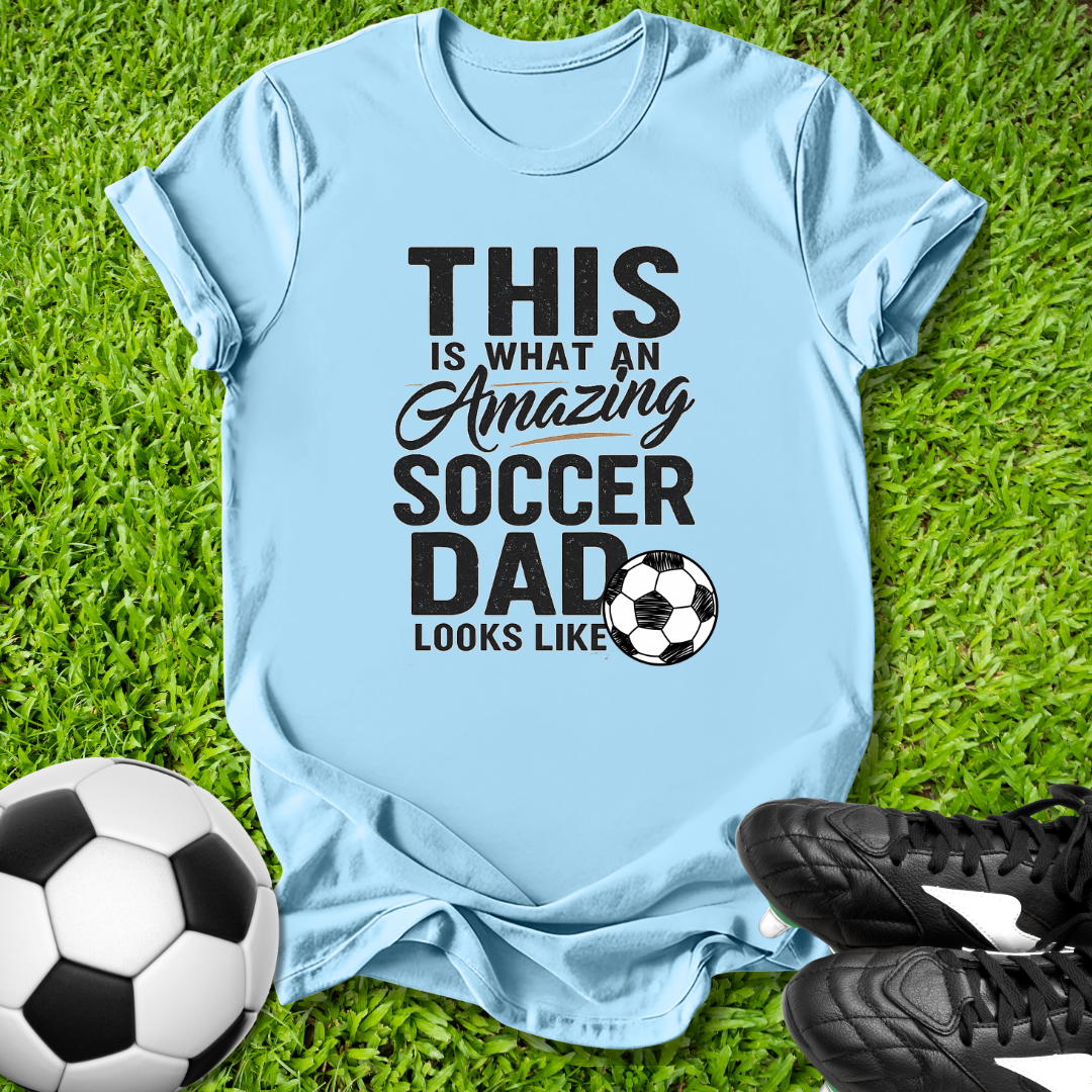 Light blue t-shirt with 'This is what an amazing soccer dad looks like' text on grass with soccer ball and shoes.