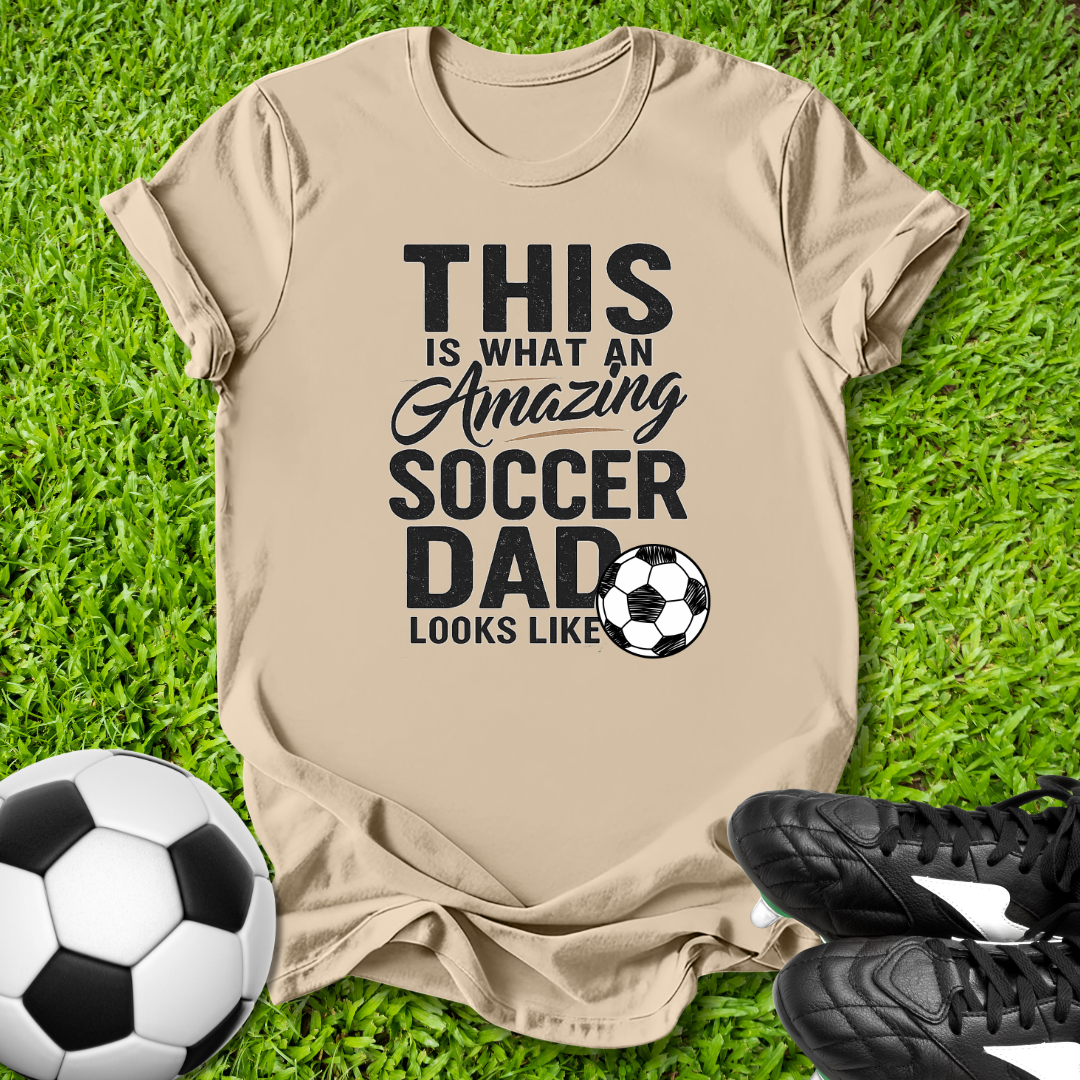 Beige t-shirt with 'This is what an amazing soccer dad looks like' text on grass with soccer ball and shoes.