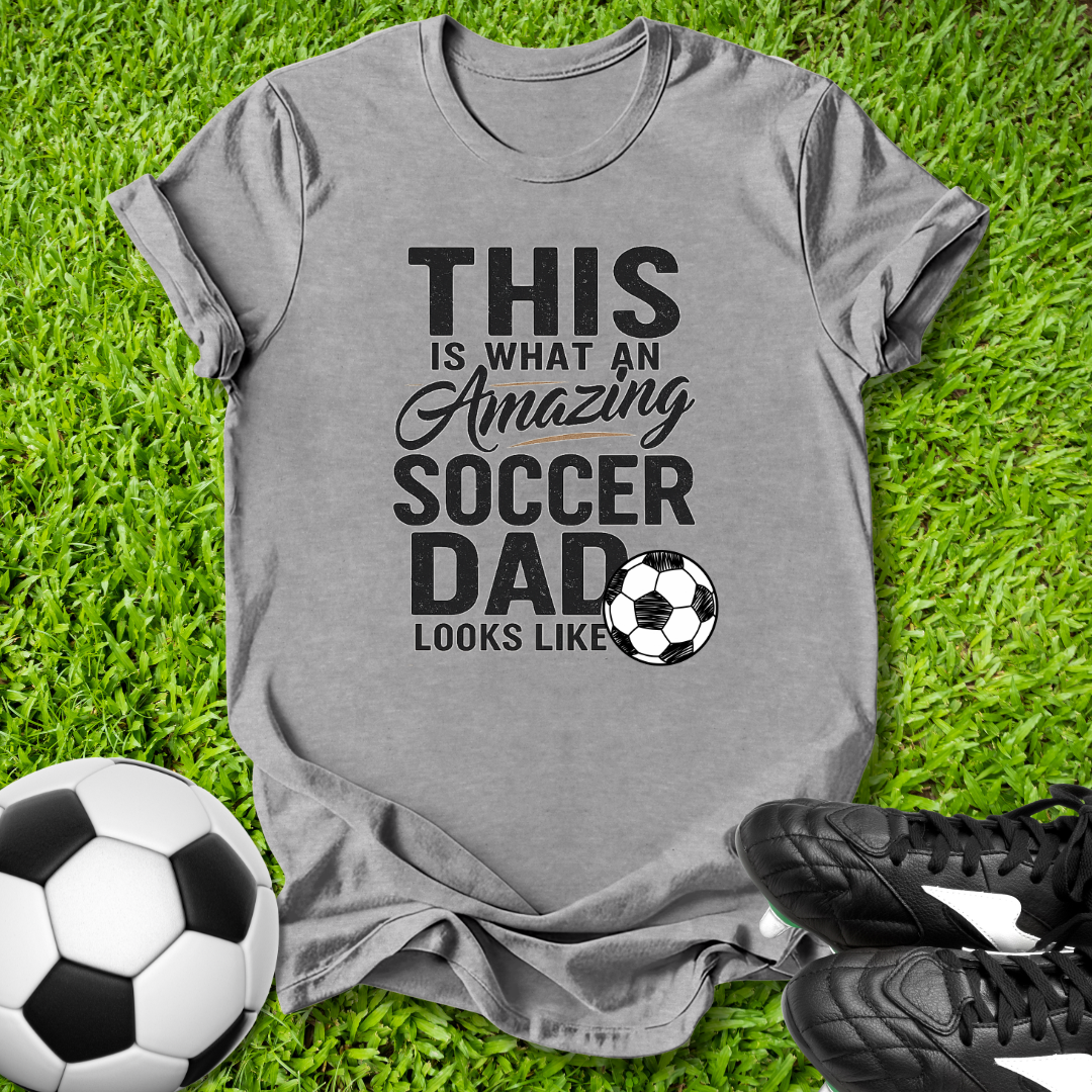 Gray t-shirt with 'This is what an amazing soccer dad looks like' text on grass with soccer ball and shoes.