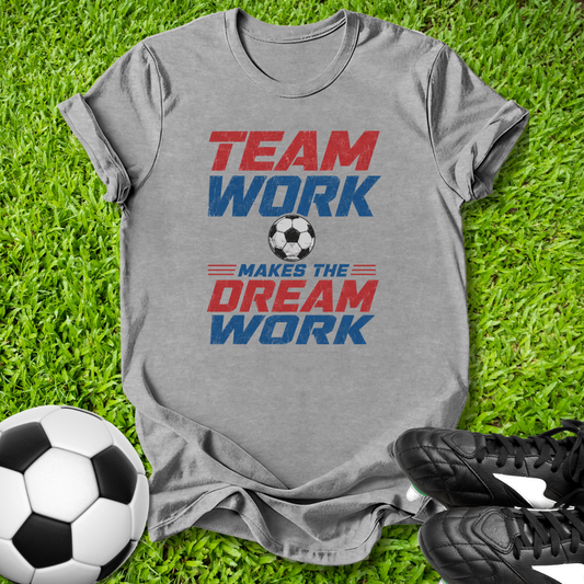 Gray t-shirt with 'Team Work Makes the Dream Work' text on grass with soccer ball and shoes.