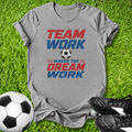Gray t-shirt with 'Team Work Makes the Dream Work' text on grass with soccer ball and shoes.