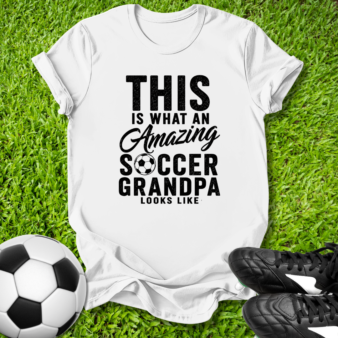 White t-shirt with 'Amazing Soccer Grandpa' text on grass with soccer ball and shoes