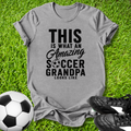 Gray t-shirt with 'Amazing Soccer Grandpa' text on grass with soccer ball and shoes.