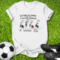 White t-shirt with soccer-themed graphic on grass with soccer ball and shoes