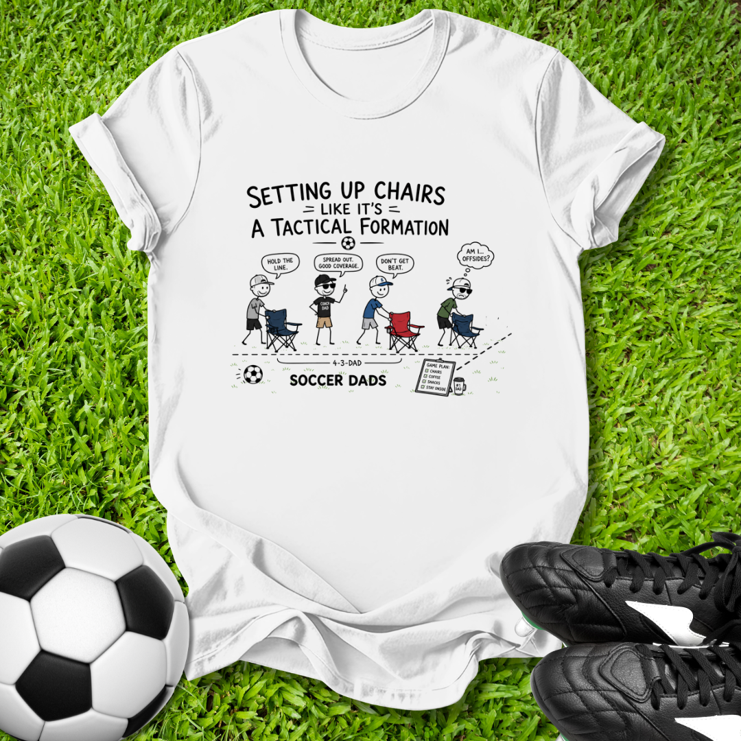 White t-shirt with soccer-themed graphic on grass with soccer ball and shoes