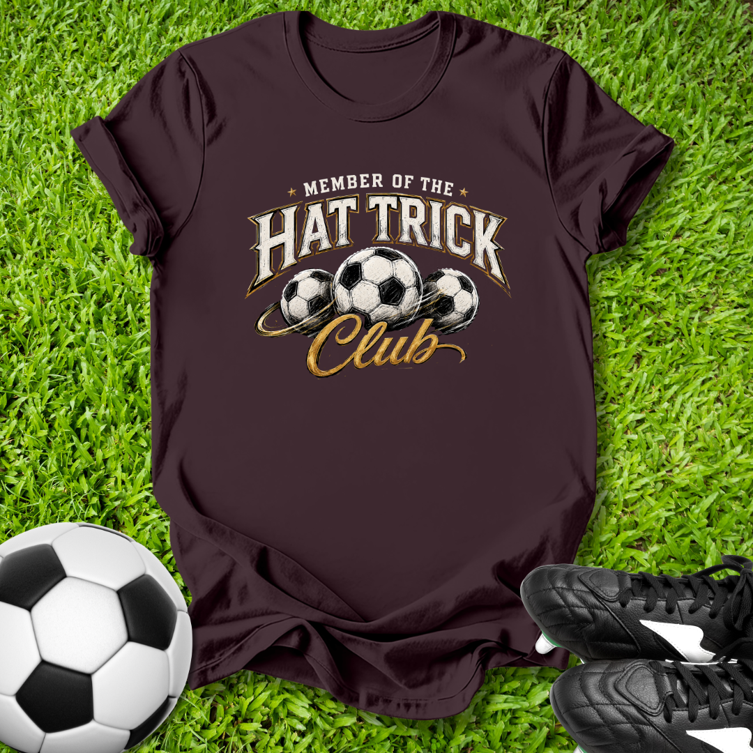 Maroon t-shirt with 'Member of the Hat Trick Club' text and soccer ball graphics on grass with soccer ball and shoes.