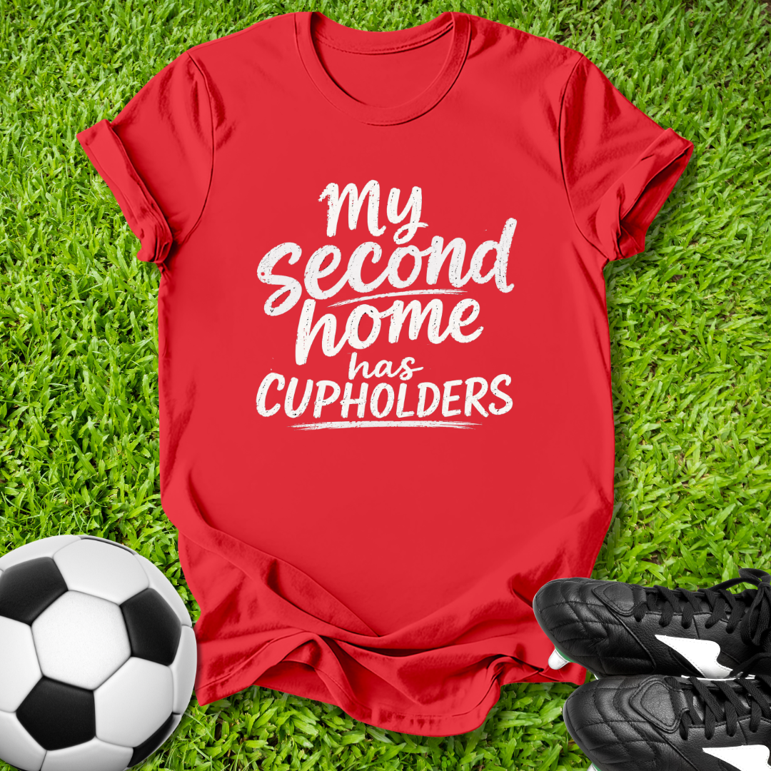 Red t-shirt with 'My second home has cupholders' text on grass with soccer ball and shoes.