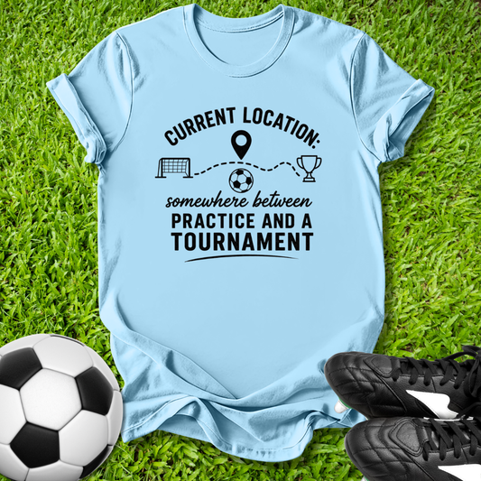 Light blue t-shirt with soccer-themed text on grass with soccer ball and shoes