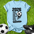Light blue t-shirt with soccer-themed design on grass with soccer ball and shoes.
