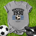 Gray t-shirt with 'Montevideo 1930' design on grass with soccer balls and shoes.