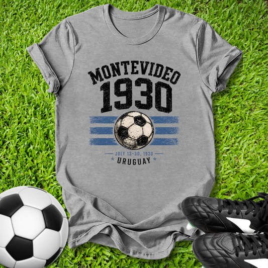 Gray t-shirt with 'Montevideo 1930' design on grass with soccer balls and shoes.