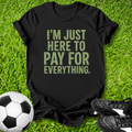 Black t-shirt with 'I'm just here to pay for everything' text on grass with soccer ball and shoes.