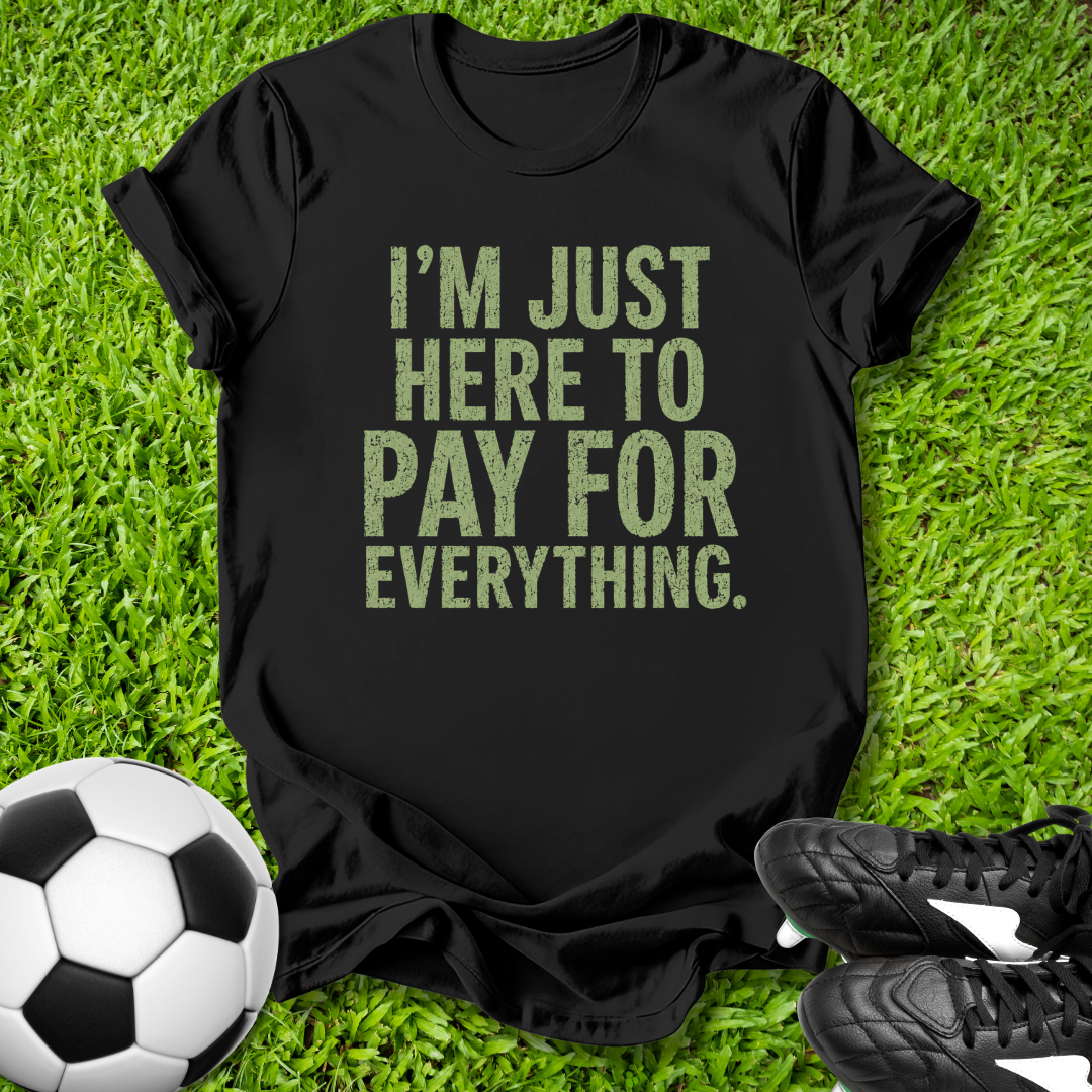 Black t-shirt with 'I'm just here to pay for everything' text on grass with soccer ball and shoes.