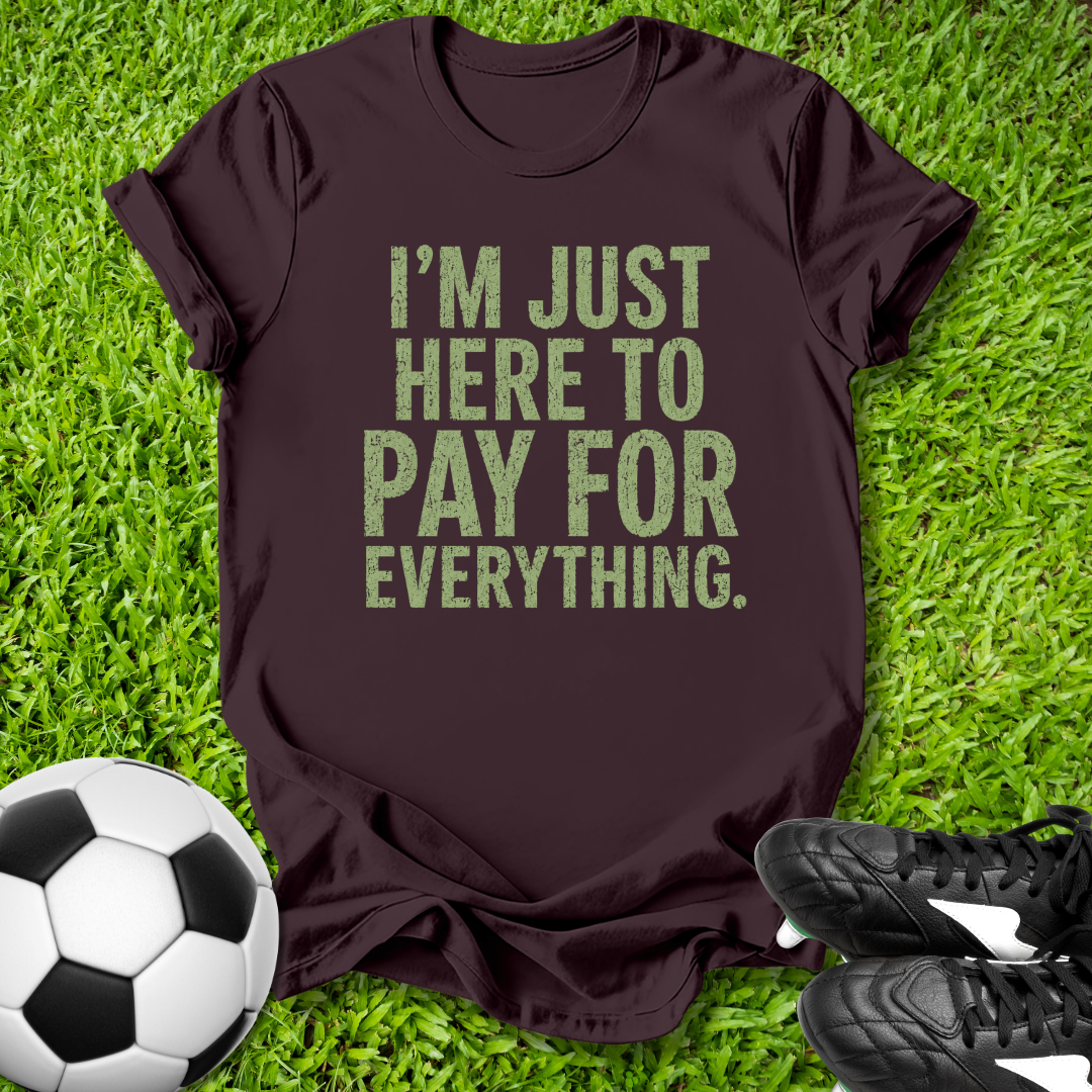 Brown t-shirt with 'I'm just here to pay for everything' text on grass with soccer ball and shoes.