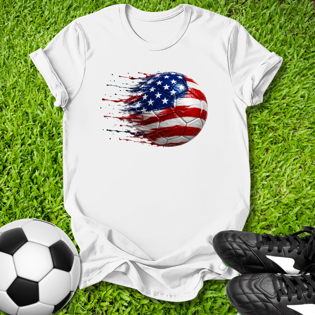 White t-shirt with American flag soccer ball design on grass with soccer ball and shoes.