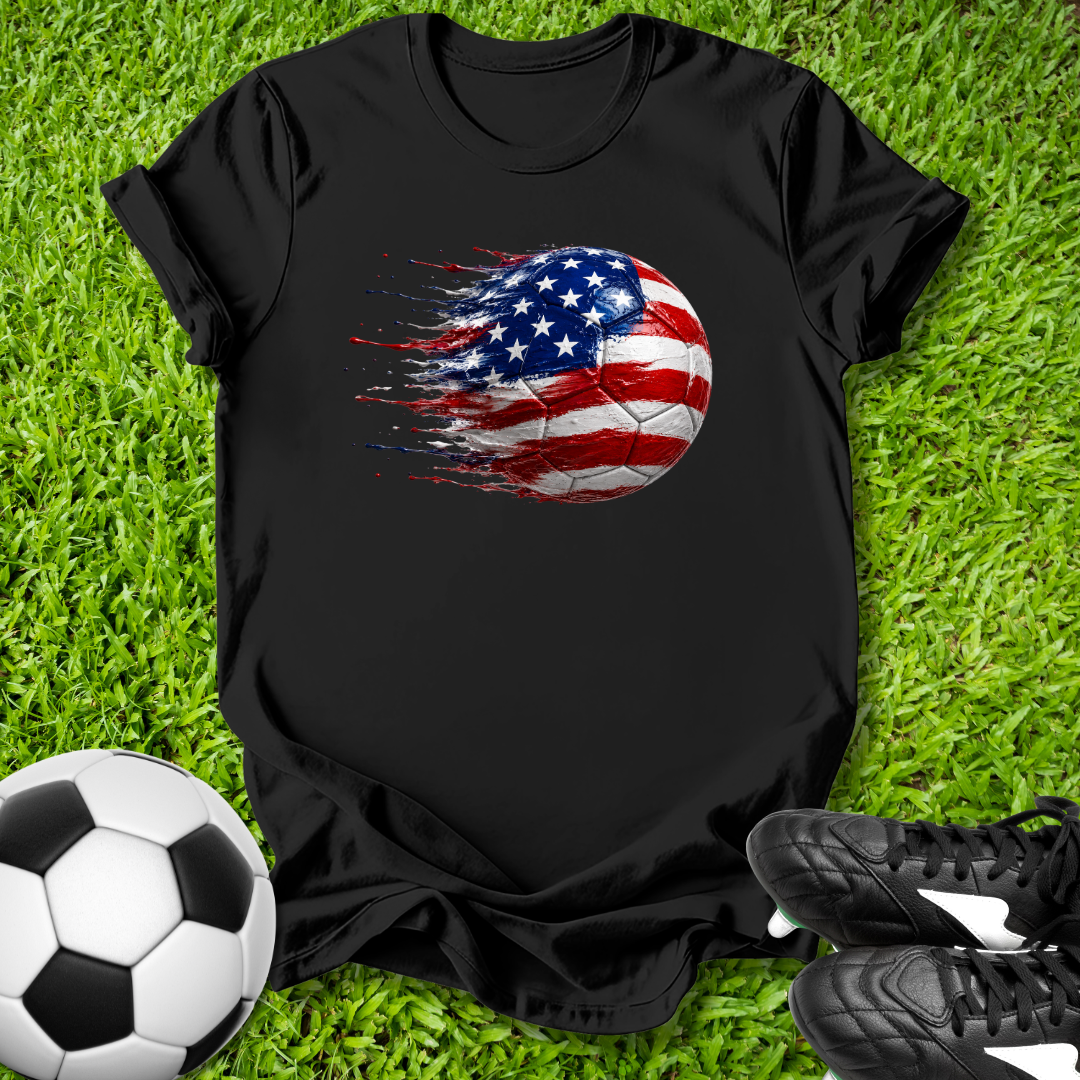Black t-shirt with American flag soccer ball design on grass with soccer ball and shoes.