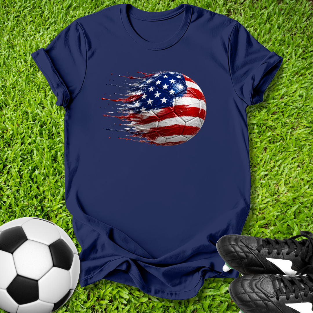 Navy blue t-shirt with American flag soccer ball design on grass with soccer ball and shoes.