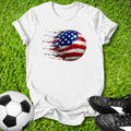 White t-shirt with American flag soccer ball design on grass with soccer ball and shoes.