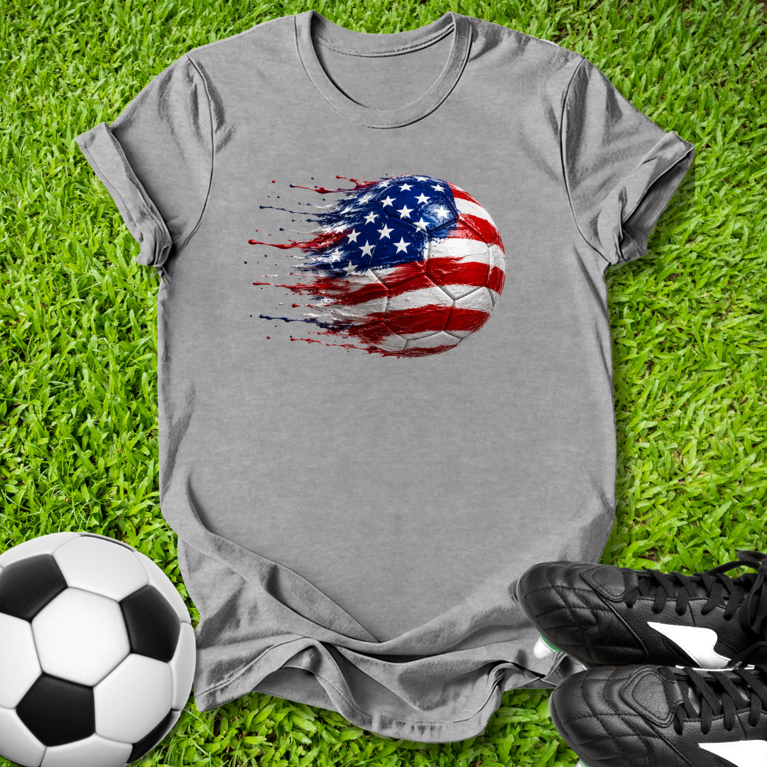 Gray t-shirt with American flag soccer ball design on grass with soccer ball and shoes.