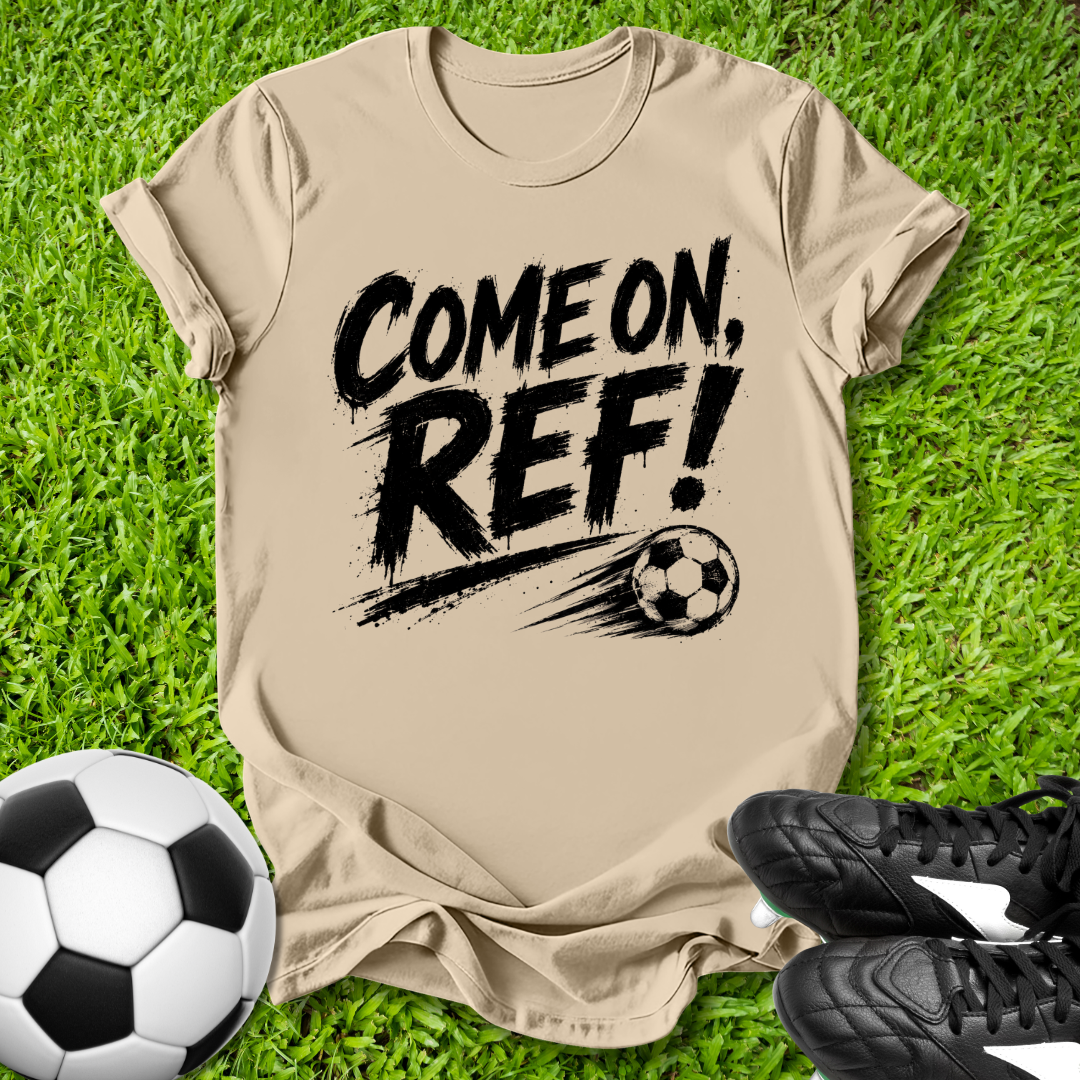 Beige t-shirt with 'COME ON, REF!' text on grass with soccer ball and shoes.