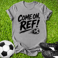 Gray t-shirt with 'COME ON, REF!' text on grass with soccer balls and shoes.
