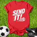 Red t-shirt with 'SEND IT!' and soccer ball graphic on grass with soccer ball and shoes.