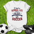 White t-shirt with soccer-themed text on grass with soccer ball and shoes