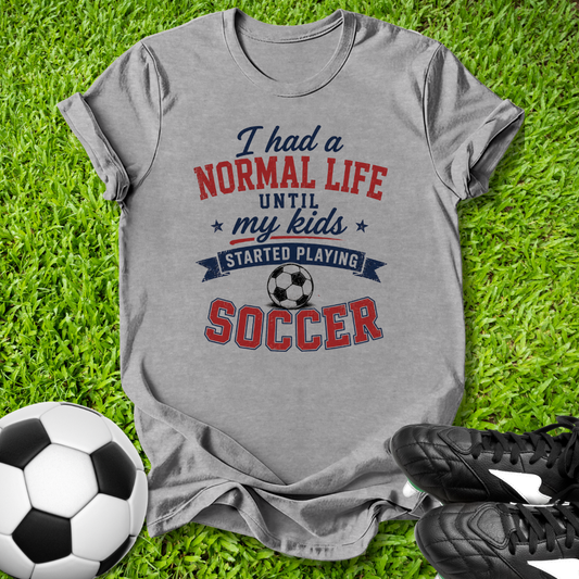 Gray t-shirt with soccer-themed text on grass with soccer ball and shoes