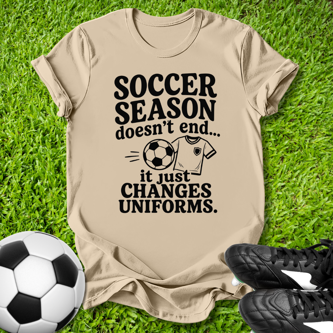 Beige t-shirt with 'Soccer Season doesn't end...' text on grass with soccer ball and shoes.