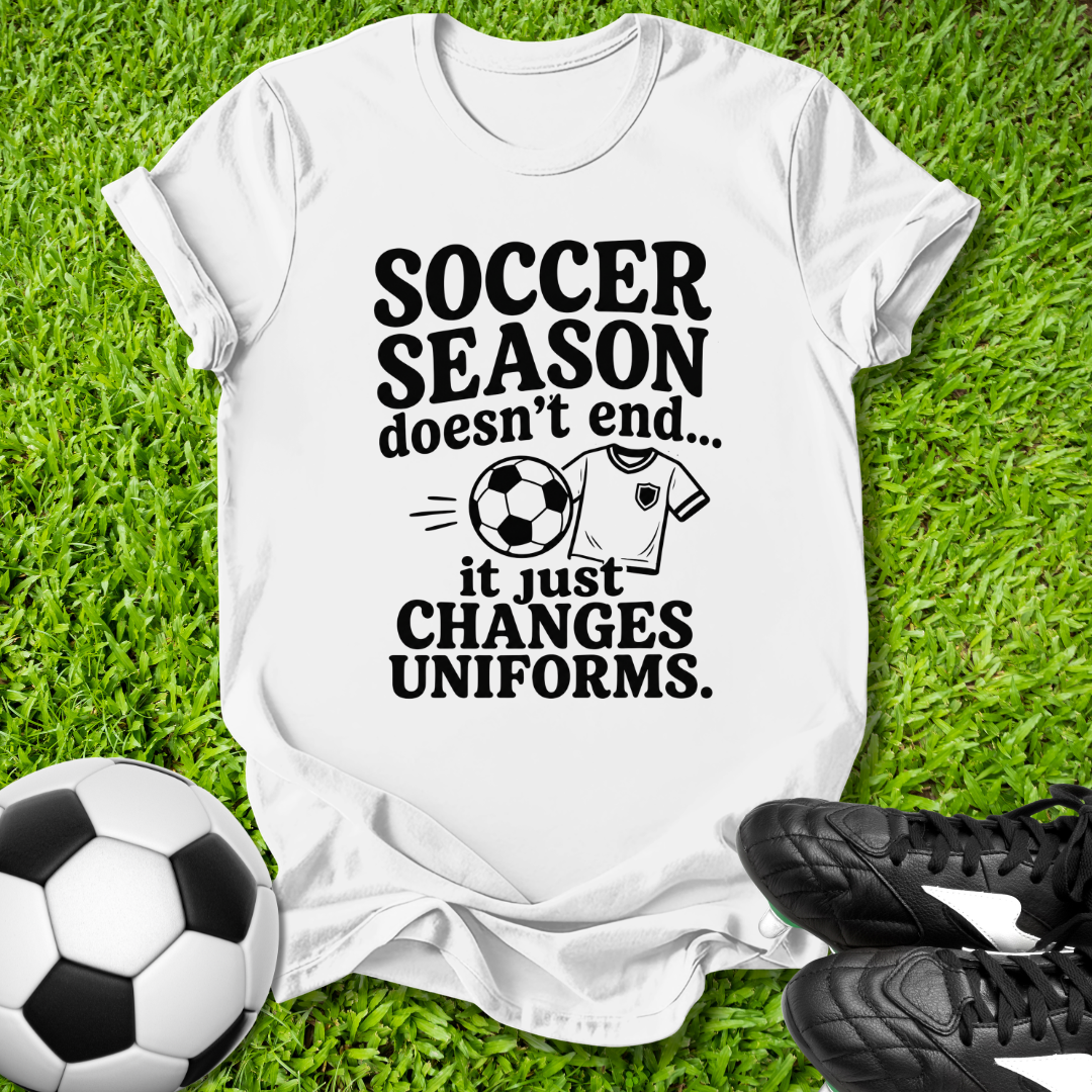 White t-shirt with soccer-themed text on grass with soccer ball and shoes
