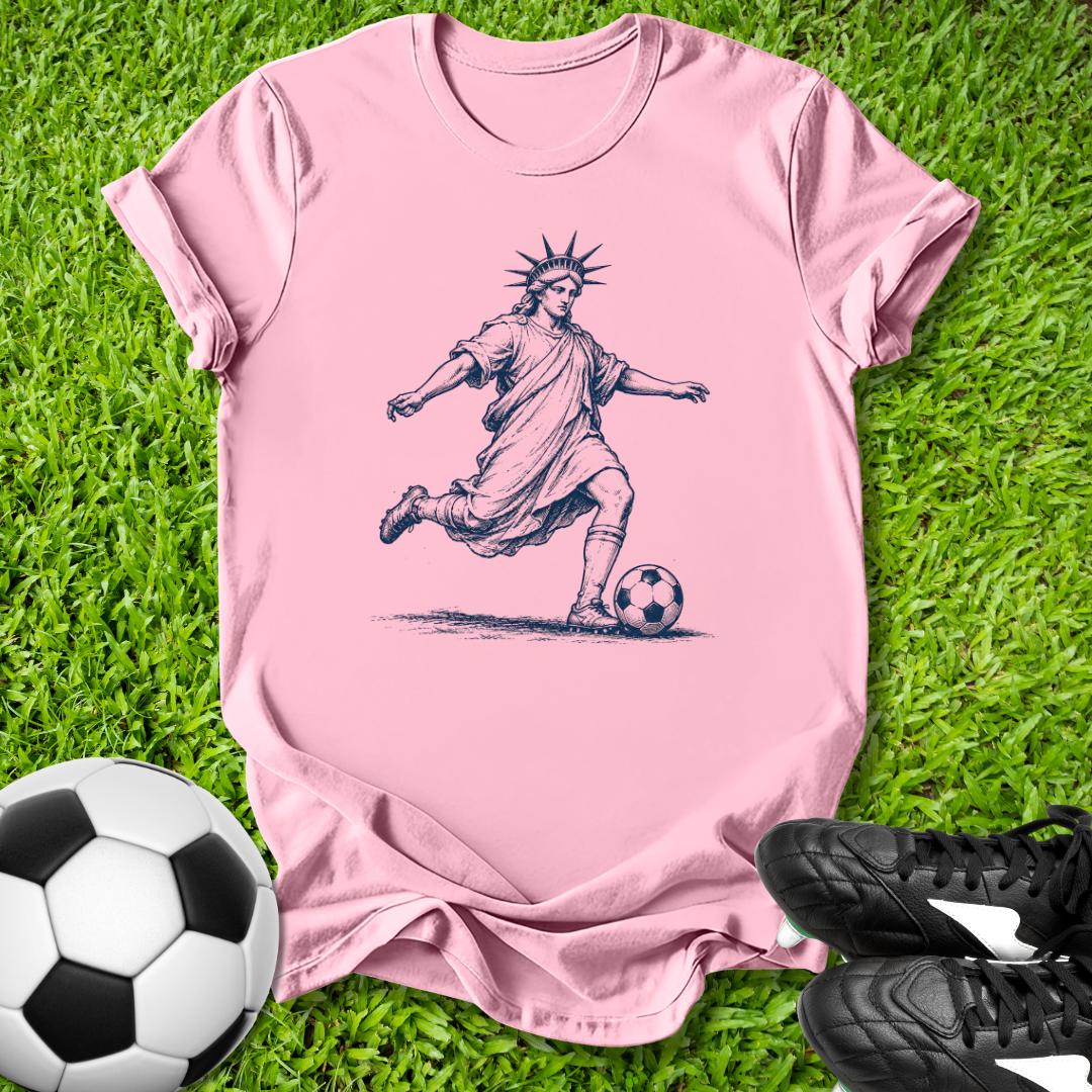 Pink t-shirt with a graphic of a soccer player resembling the Statue of Liberty on grass with soccer balls and shoes.