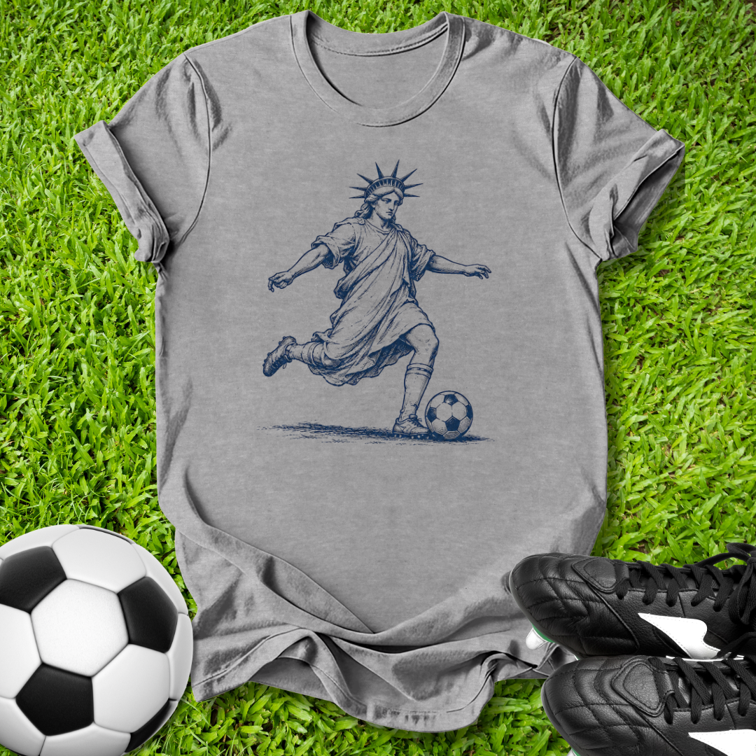 Gray t-shirt with a graphic of the Statue of Liberty playing soccer on grass with soccer ball and shoes.