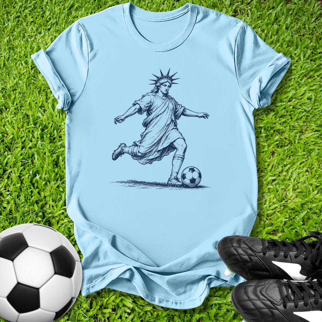 Light blue t-shirt with a graphic of the Statue of Liberty playing soccer on grass with soccer ball and shoes.