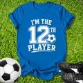 Blue t-shirt with 'I'm the 12th Player' text on grass with soccer ball and shoes.