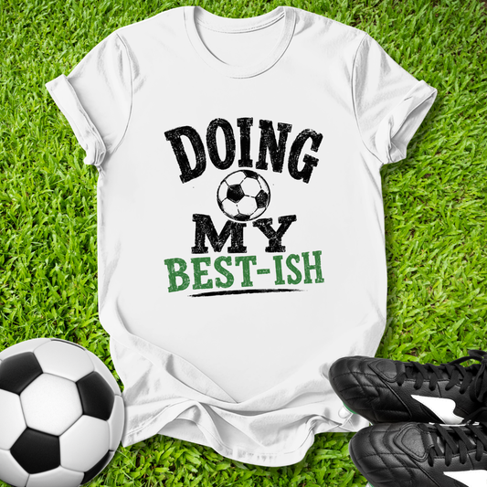 White t-shirt with 'Doing My Best-ish' text on grass with soccer ball and shoes