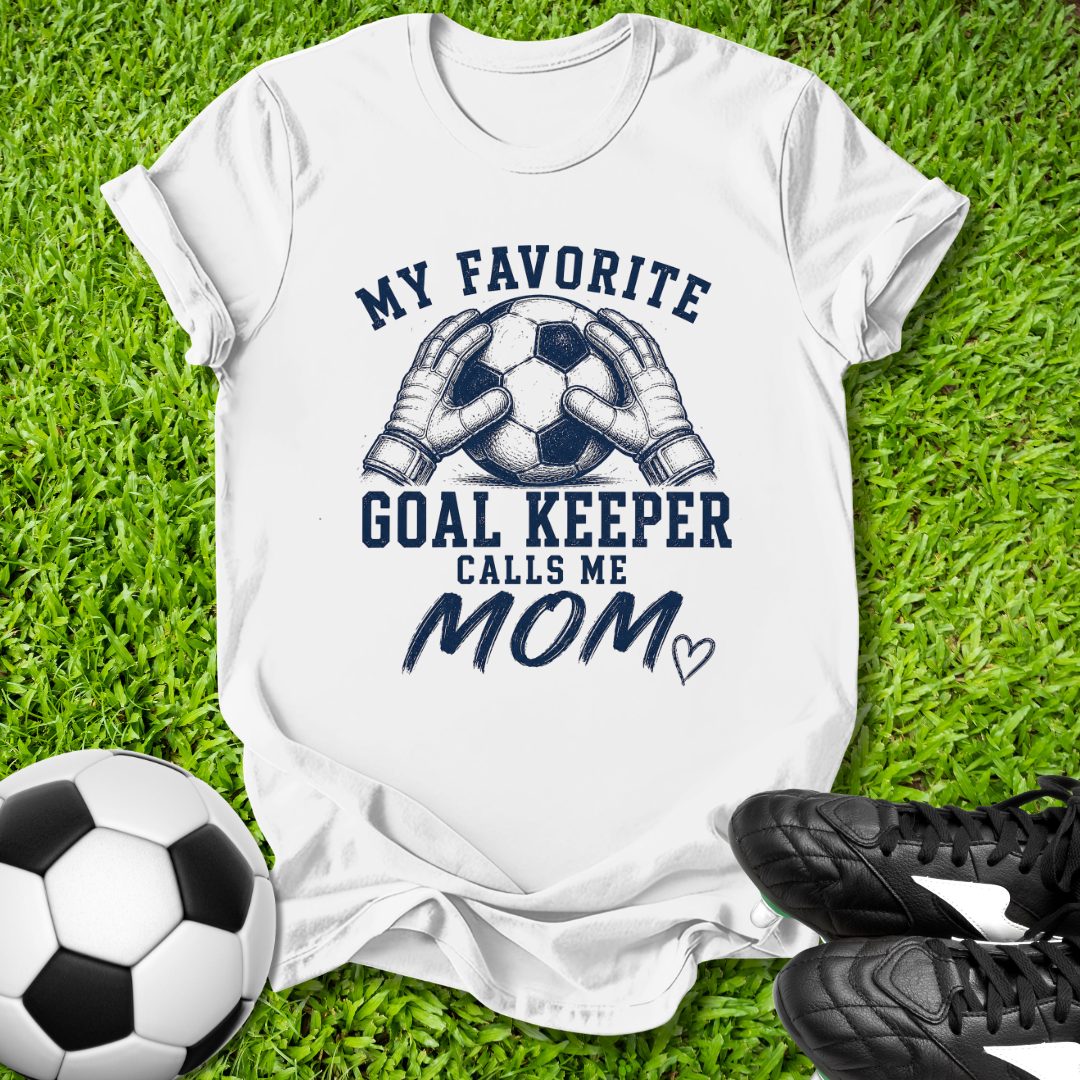 White t-shirt with 'My Favorite Goal Keeper Calls Me Mom' text on grass with soccer ball and shoes.