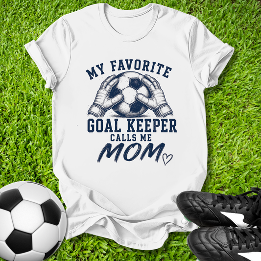 White t-shirt with 'My Favorite Goal Keeper Calls Me Mom' text on grass with soccer ball and shoes.