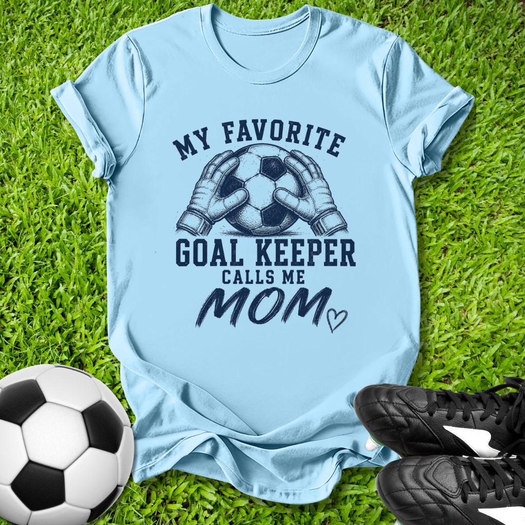 Light blue t-shirt with soccer-themed design on grass with soccer ball and shoes.