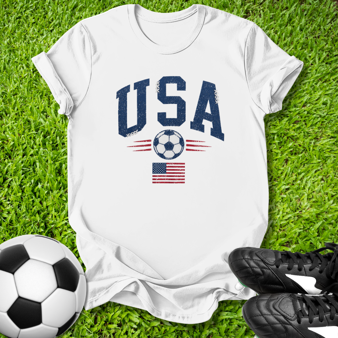 White t-shirt with 'USA' and soccer ball design on grass with soccer ball and shoes.