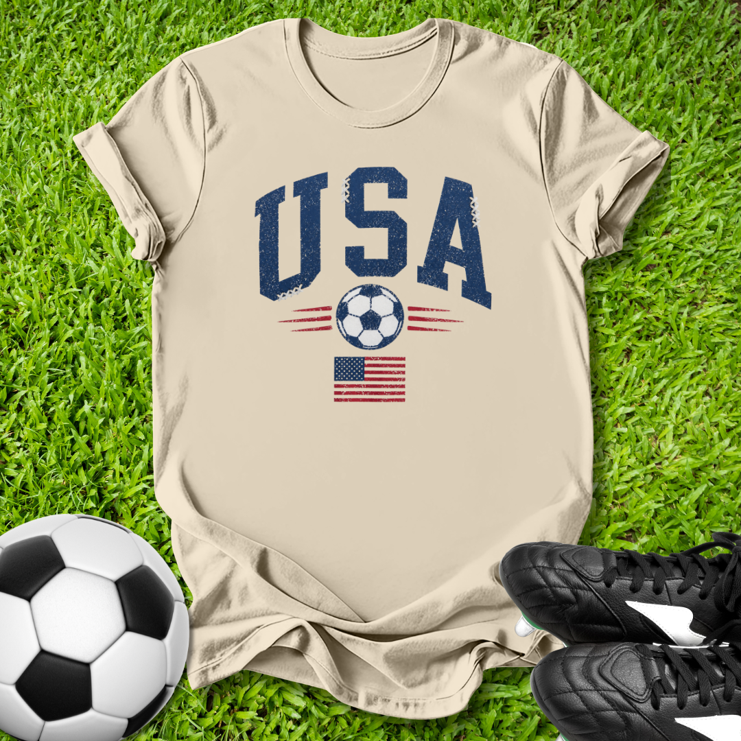 Beige t-shirt with 'USA' and soccer ball design on grass with soccer ball and shoes.