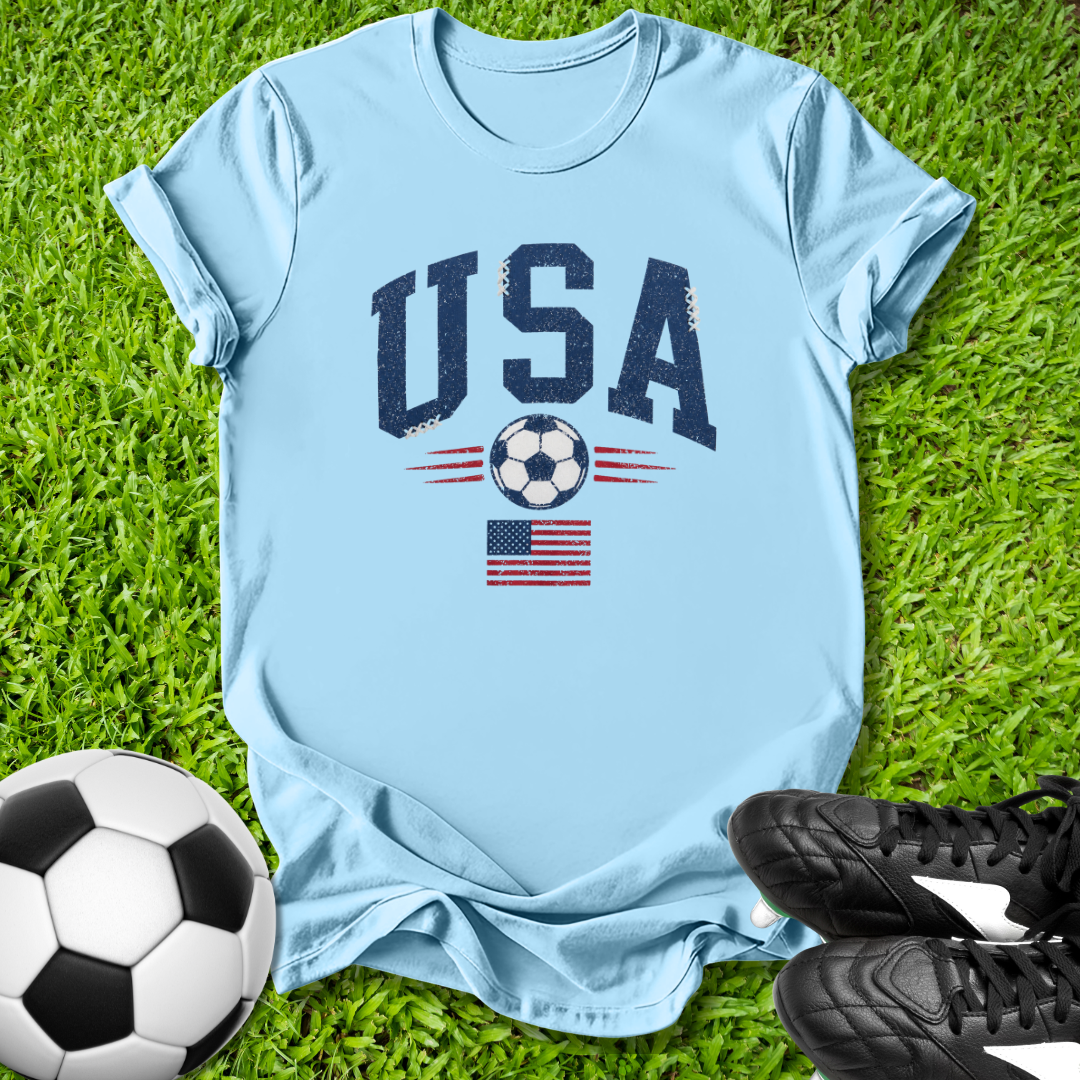 Gray t-shirt with 'USA' and soccer ball design on grass with soccer ball and shoes.