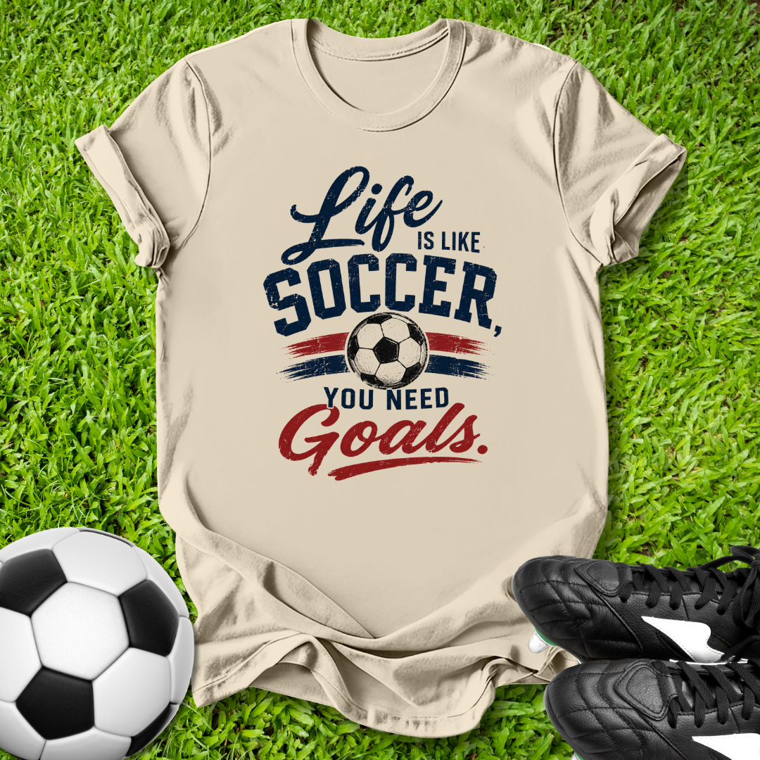 Beige t-shirt with 'Life is like soccer, you need goals' text on grass with soccer ball and shoes.