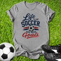 Gray t-shirt with 'Life is like Soccer, you need Goals' text on grass with soccer ball and shoes.