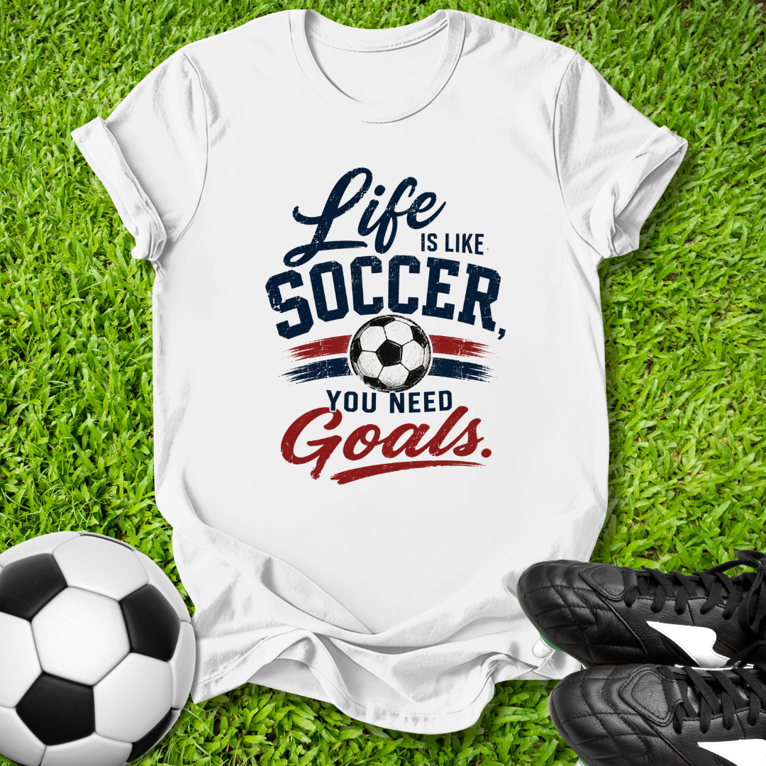 White t-shirt with 'Life is like soccer, you need goals' text on grass with soccer ball and shoes.