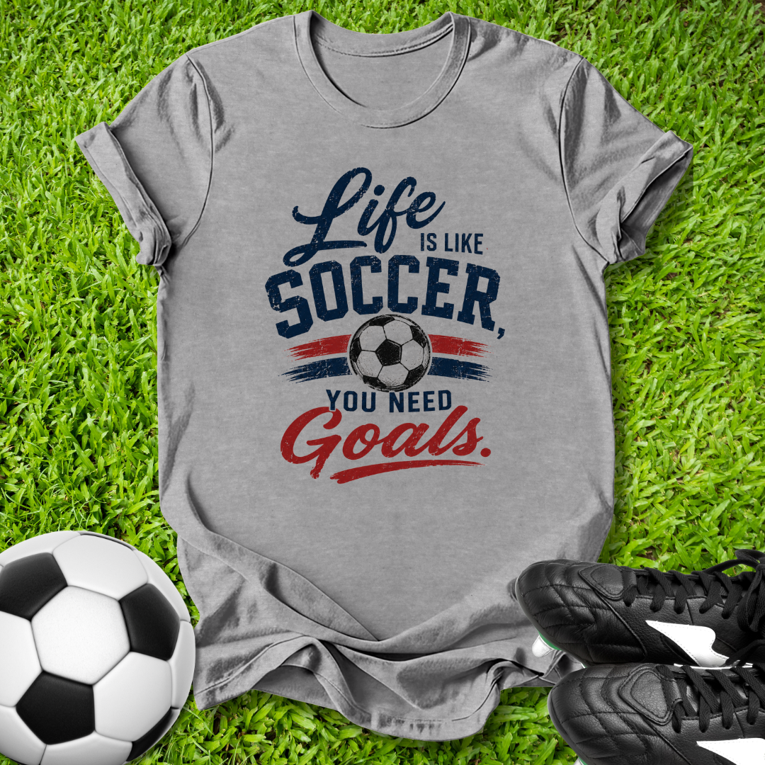 Gray t-shirt with 'Life is like Soccer, you need Goals' text on grass with soccer ball and shoes.