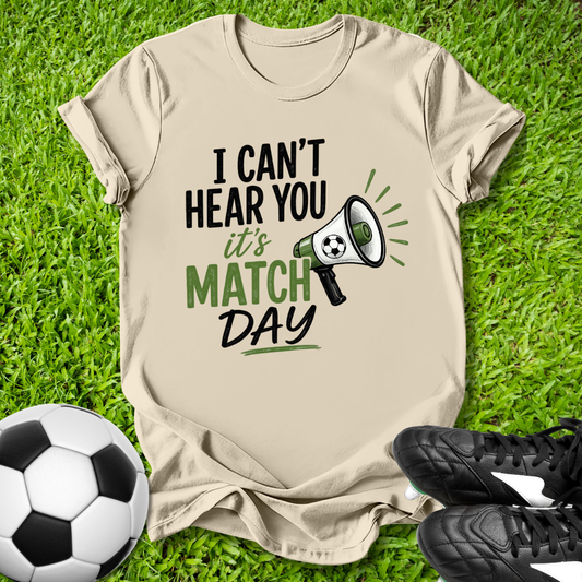 Beige t-shirt with 'I can't hear you, it's match day' text on grass with soccer ball and shoes.