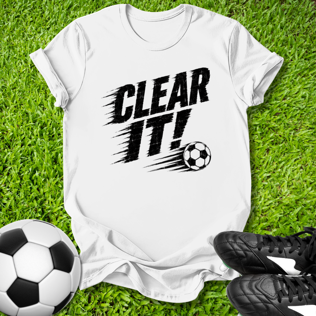 White t-shirt with 'CLEAR IT!' text and soccer ball graphic on grass with soccer ball and shoes.