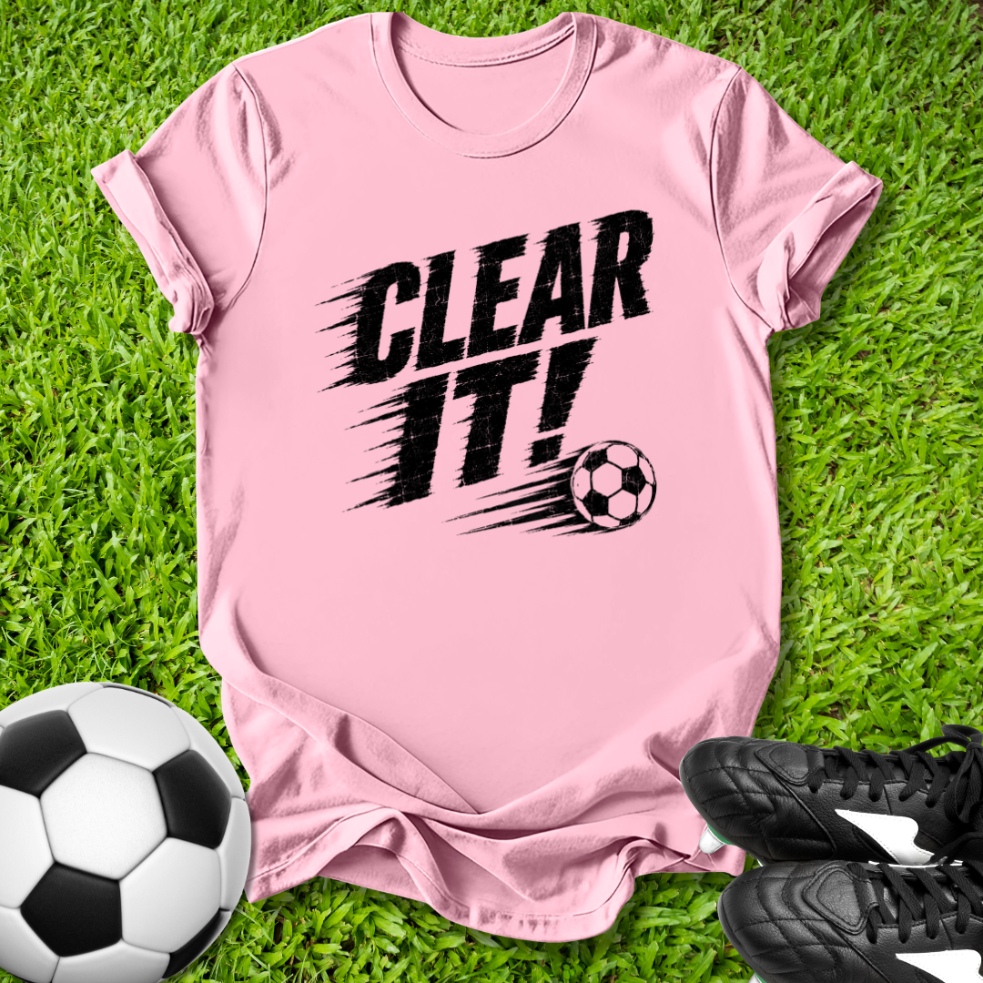 Pink t-shirt with 'CLEAR IT!' text and soccer ball on grass with soccer ball and shoes.