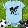 Light blue t-shirt with 'CLEAR IT!' text and soccer ball graphic on grass with soccer balls and shoes.