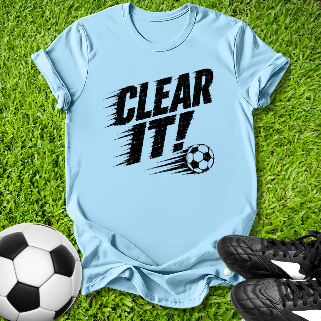 Light blue t-shirt with 'CLEAR IT!' text and soccer ball graphic on grass with soccer balls and shoes.